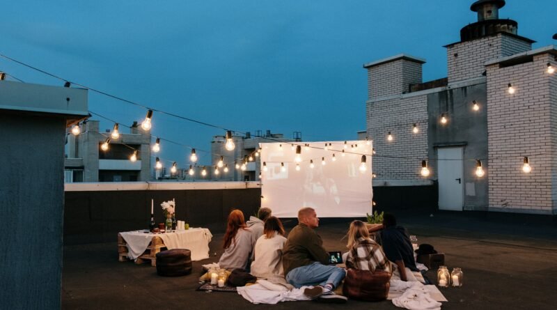 Friends watching an outdoor movie on a rooftop at dusk under string lights