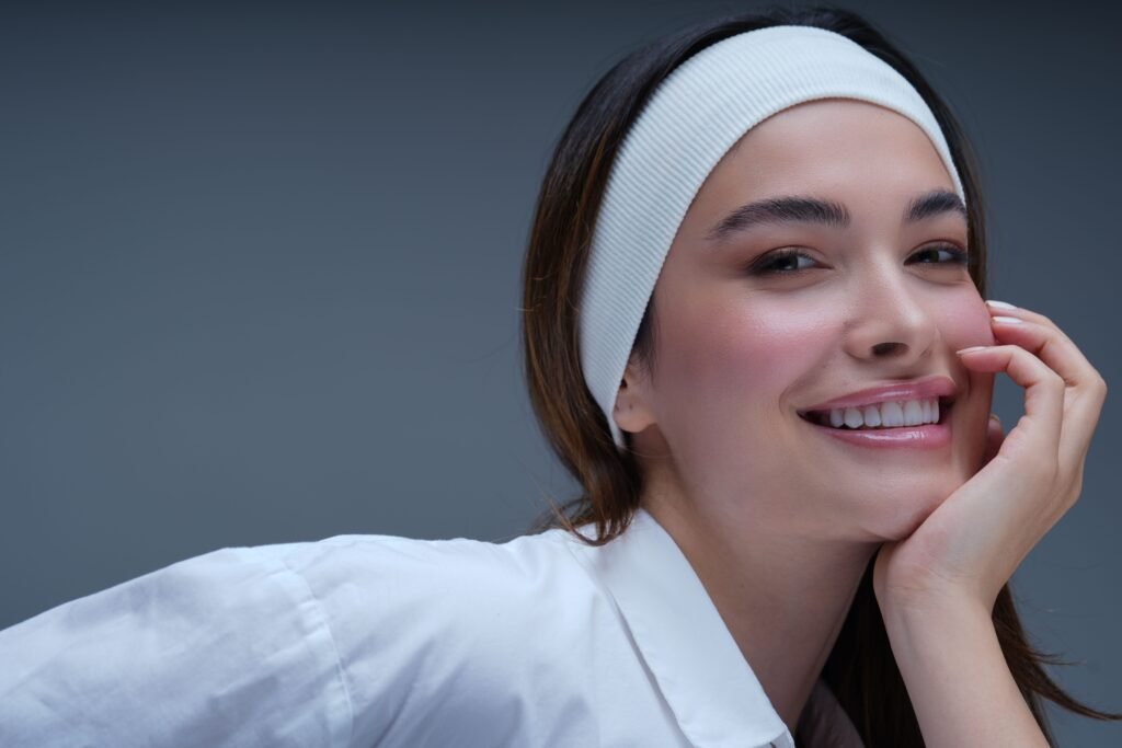 Smiling young woman with a white headband and glowing dewy skin, resting her face on her hand