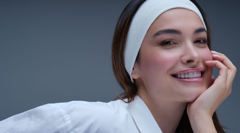 Smiling young woman with a white headband and glowing dewy skin, resting her face on her hand