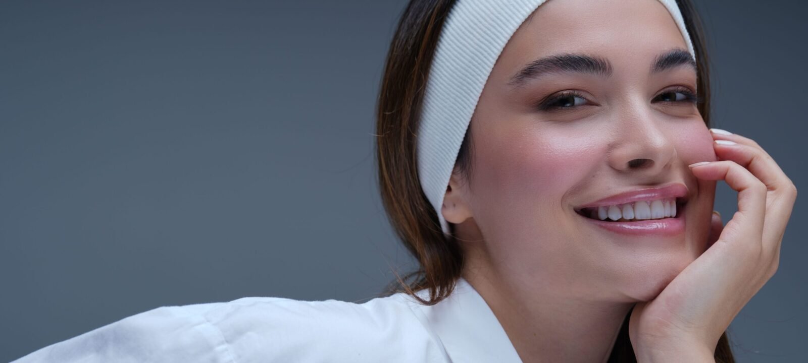 Smiling young woman with a white headband and glowing dewy skin, resting her face on her hand
